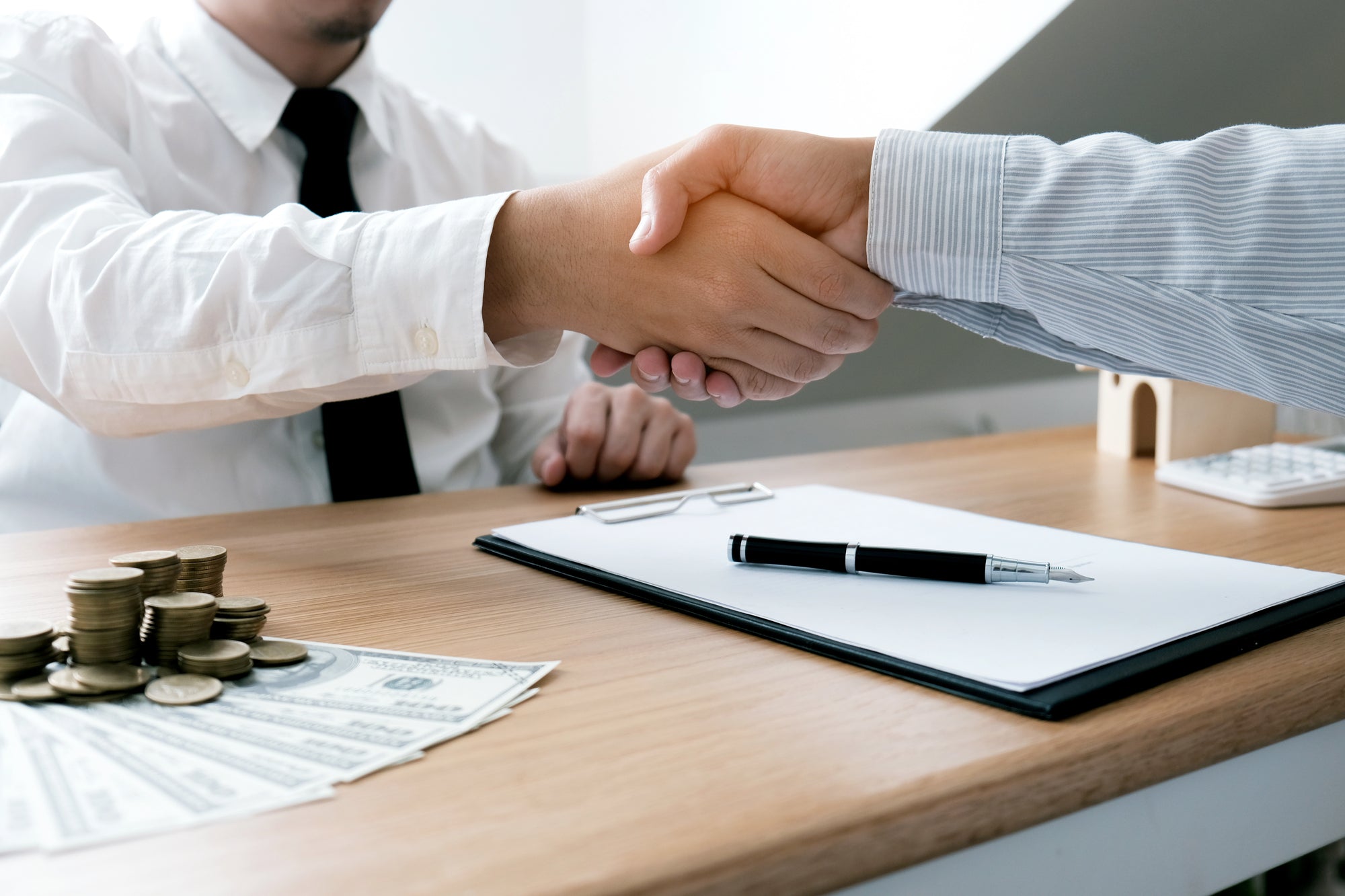Two people shaking hands over a desk with money, coins, and documents.